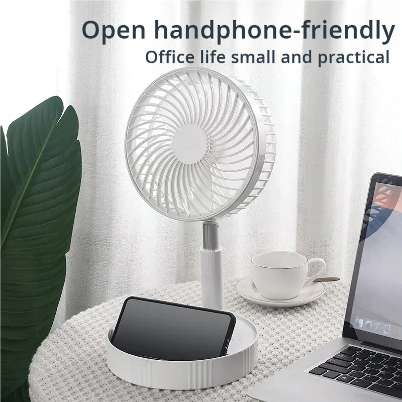 Small white fan on a desk with a phone, laptop, and cup in an office setting.